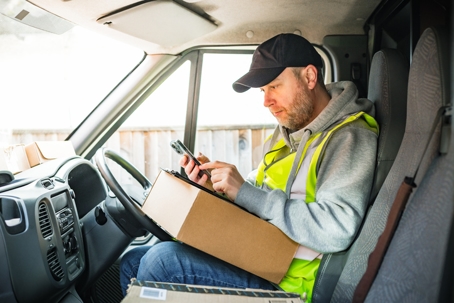 A delivery driver in a high-visibility vest sits in a van, holding a package and using a smartphone. | Law Offices of Steven J. Klearman & Associates