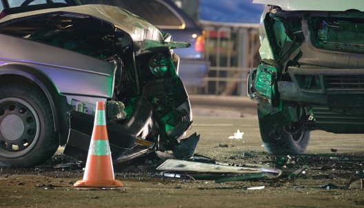 Two heavily damaged vehicles after a nighttime car accident, with debris on the road and an orange traffic cone in the foreground. | Law Offices of Steven J. Klearman & Associates